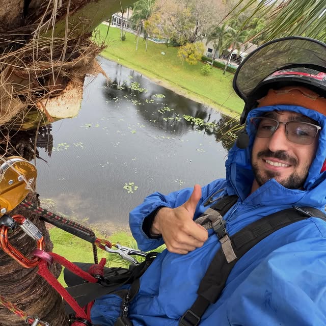 Fadi from Elannan Tree Service, smiling, harnessed in a palm tree, with a Boca-area pond and homes behind him